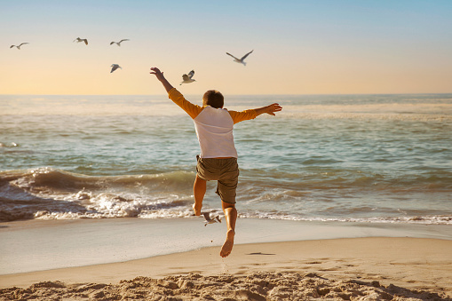 Young boy jumping for joy at the beach during sunset with birds flying