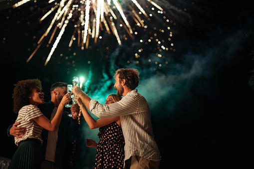 Four friends standing outside, toasting with glasses of wine and watching fireworks