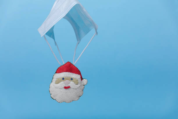 istock Santa Claus in a red hat is on protective face mask shaped  parachute levitate on blue background. Quarantine celebration, Pandemic Christmas. Covid-19 holidays. New normal. 1284961320