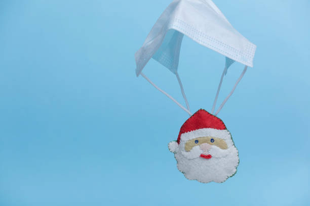 istock Santa Claus in a red hat is on protective face mask shaped  parachute levitate on blue background. Quarantine celebration, Pandemic Christmas. Covid-19 holidays. New normal. 1284961329