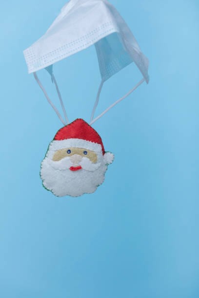 istock Santa Claus in a red hat is on protective face mask shaped  parachute levitate on blue background. Quarantine celebration, Pandemic Christmas. Covid-19 holidays. New normal. 1284961331