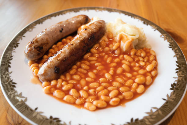Two large fried sausages with baked beans and mashed potato on a white plate with a gold colour trim on a wooden table lit by window natural light.
