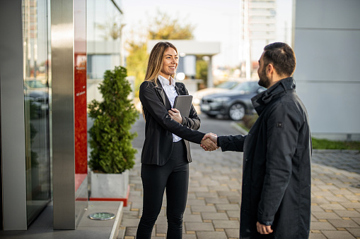 Young man arriving at car dealership while saleswoman welcoming him at entrance and greeting with handshake