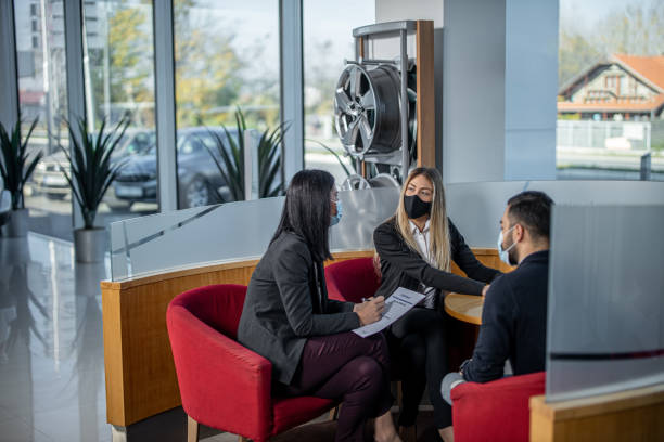 Two female colleagues sitting and wearing face mask with male customer in car dealership Two young female colleagues sitting and wearing face mask with male customer in car dealership and talking car salesperson photos stock pictures, royalty-free photos & images