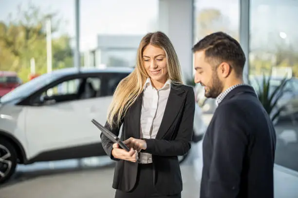 Smiling car saleswoman standing and showing to male customer car performance on tablet in saloon