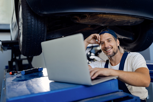 Happy car mechanic using laptop while working at auto repair shop and looking at camera.