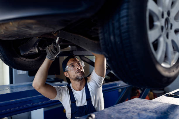 Car mechanic repairing chassis of a vehicle in while working in a workshop. Mid adult mechanic repairing undercarriage of a car in auto repair shop. mechanic working under car stock pictures, royalty-free photos & images