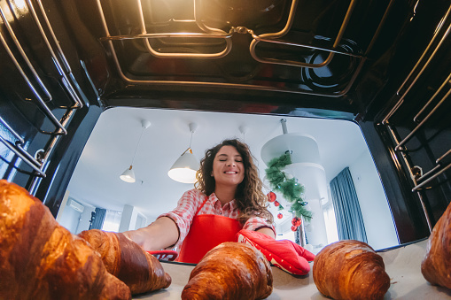 Young woman leaning towards the open oven and taking a baking pan with croissants out of it