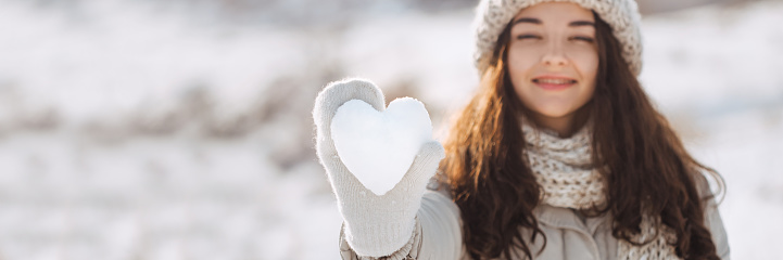 Snow heart in woman's hand outdoors. Girl on stylish winter clothes hat, warm beige gloves holding snowy heart against a nature winter background. I love winter or St.Valentine's Day romantic creative concept. Mixed race Asian Caucasian model. Panorama banner with free space for text mockup.