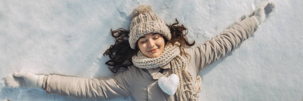 Portrait of a beautiful young woman laying down on a frozen snow lake playing games during a sunny winter vacation. Mixed race Asian Caucasian gorgeous girl resting during winter holidays. Snowball in the shape of the heart on woman's chest. Panorama banner with free space for text mockup.