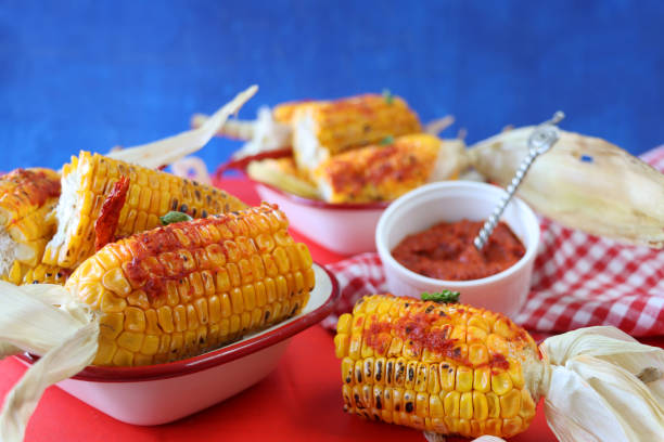 istock Image of enamel bowls on red surface containing dent corn variety corn on the cob with peeled back husks spread with chili sauce, bowl of red Mexican chili sauce, blue background, focus on foreground 1284978243