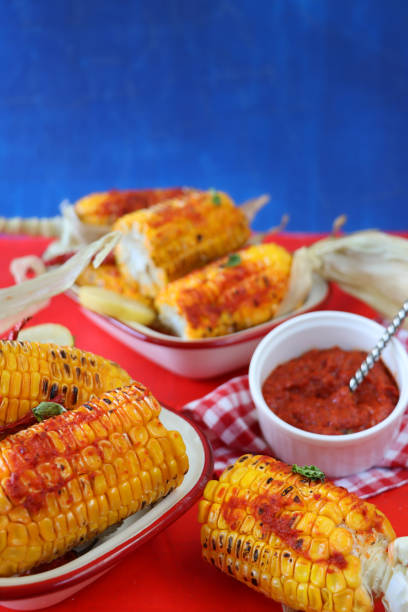 istock Image of enamel bowls on red surface containing dent corn variety corn on the cob with peeled back husks spread with chili sauce, bowl of red Mexican chili sauce, blue background, focus on foreground 1284978335