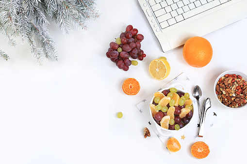 Modern woman work desk, healthy breakfast, home office. Computer, Christmas decorations, fruit salad and a cup of tea on a light table. Lifestyle concept, flat lay, holidays eve