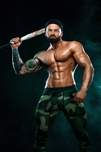 istock Strong and fit man bodybuilder with baseball bat shows abdominal muscles under a t-shirt. Sporty muscular guy athlete. Sport and fitness concept. Men's fashion. 1284983317