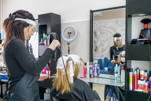 Hair stylist with visor on her face blow drying colored hair