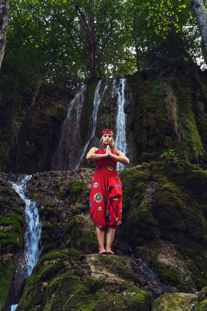 vista anteriore ritratto di donna caucasica adulta 30 anni sulla roccia in natura di fronte alla cascata in abito rosso in piedi in posa yoga pregare - spirituality concetto di rilassamento pace interiore a tutta lunghezza - waterfall zen like women meditating foto e immagini stock