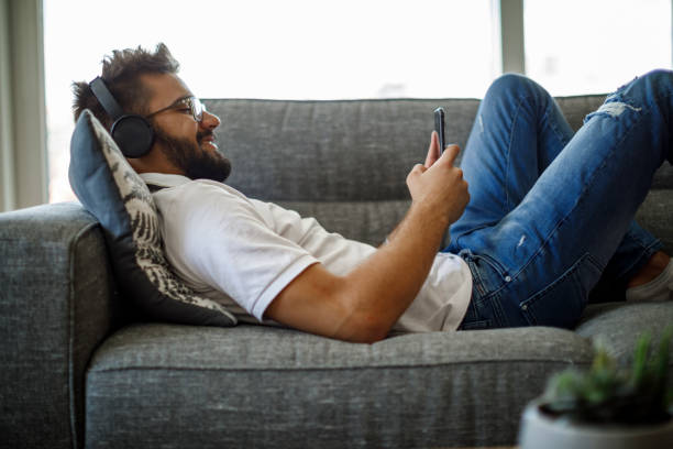 Smiling young man with wireless headphones using smart phone on sofa Smiling young man with wireless headphones using smart phone on sofa audio book stock pictures, royalty-free photos & images