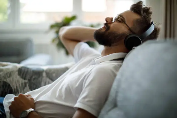 Photo of Young smiling man with bluetooth headphones relaxing on sofa