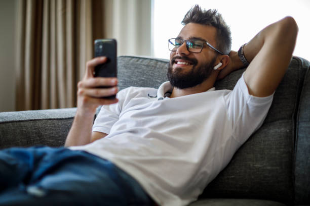 istock Smiling young man with bluetooth headphones using smart phone on sofa 1284992188