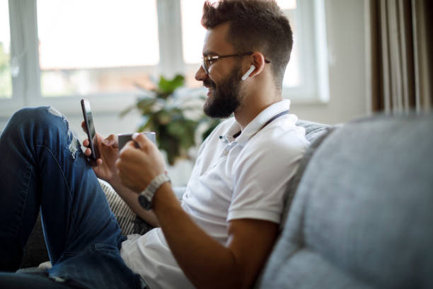 улыбающийся молодой человек с беспроводными наушниками пьет кофе и текстовые сообщения по телефону дома - sofa music teenager friendship стоковые фото и изображения