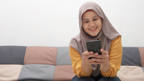 istock Beautiful Asian muslim woman using phone while sitting on sofa, girl doing chat message on smartphone and smiling 1284992305