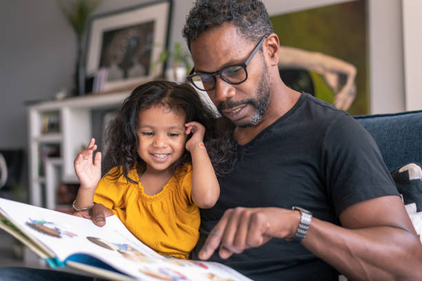 Affectionate father reading book with adorable mixed race daughter A loving father of African descent sits on the couch at home and reads a storybook to his preschool age daughter. The child is sitting on her father's lap and is smiling while looking at the book. parenting stock pictures, royalty-free photos & images