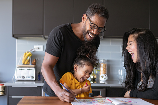 A Black man and his Asian wife laugh with their happy preschool age daughter while spending a relaxing day at home together. They are in the kitchen and the child is coloring at the table.