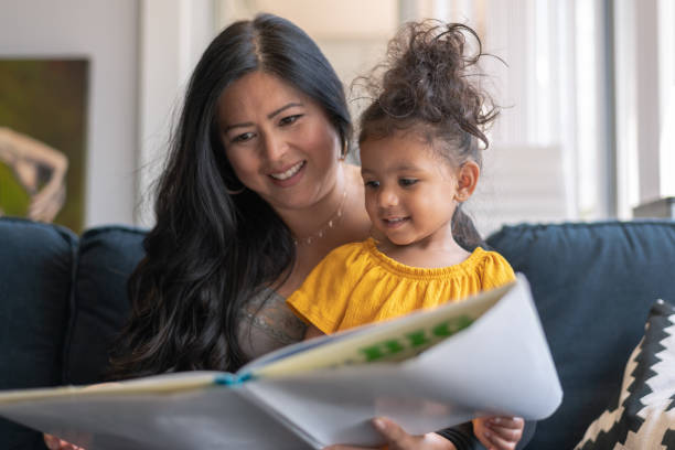 Asian mother reading book to her adorable mixed race daughter A loving mother of Asian descent sits on the couch at home and reads a storybook to her preschool age daughter. The child is sitting on her mother's lap. parent and child reading stock pictures, royalty-free photos & images