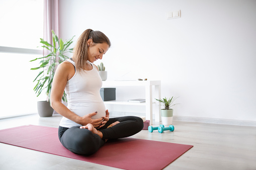 Beautiful pregnant woman sitting on exercise mat and making relaxing exercise during late pregnancy.