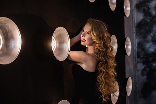 Business modern young woman in a black dress at a festive party against the background of bright ice lights. Beautiful blonde curly girl having fun at a corporate. Close up portrait. Copy space. Smile
