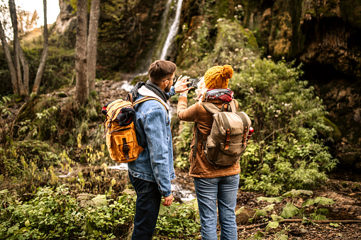 Rear view of young hiking couple with backpacks standing in a front of small forest waterfall, taking a photo