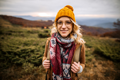 Young pretty hiker woman with knit yellow hat and blonde hair smiling, looking at camera at the top of mountain