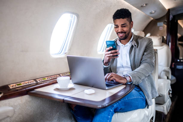 giovane uomo d'affari sorride mentre guarda il suo smartphone nel jet privato - airplane smart phone travel mobile phone foto e immagini stock