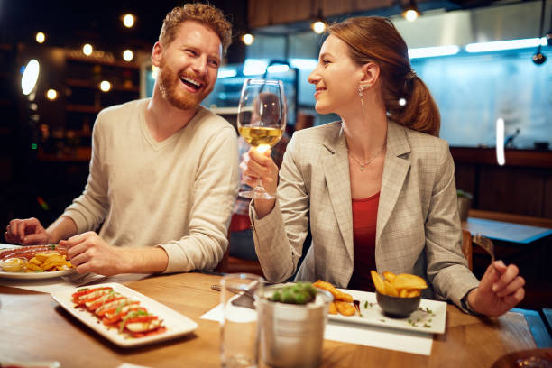 glimlachende vrolijke paarzitting in een restaurant, dat diner en het babbelen. man die aan een vrouw spreekt terwijl een vrouw die aan hem luistert en witte wijn drinkt. - dineren stockfoto's en -beelden