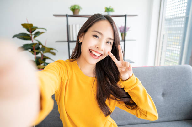 거실에 앉아 큰 미소와 예쁜 젊은 아시아 여성. 그녀는 흐린 배경에 빛 쾌활한 셀카를 찍는 재미를 가지고 - 자화상 촬영 뉴스 사진 이미지