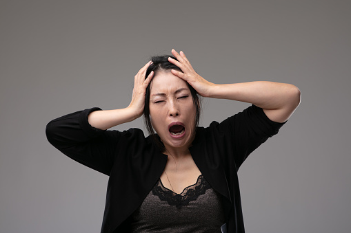 Woeful Asian woman holding her head in anguish as she cries and laments over a grey studio background with copyspace