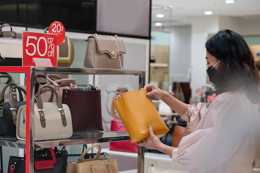 Side view shot of a Chinese woman with a face mask checking a leather handbag on sale in department store during new normal Covid 19