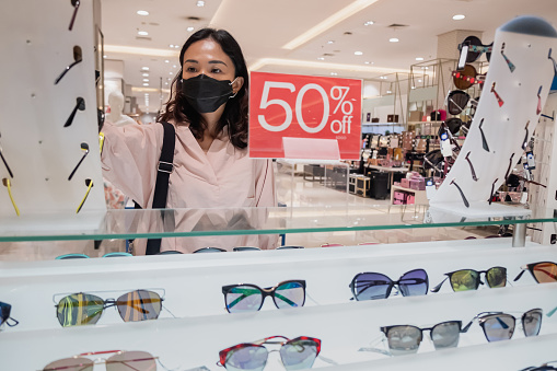 Portrait shot of an attractive Chinese woman with face mask choosing discounted sunglasses to buy in department store , amidst post pandemic Covid 19