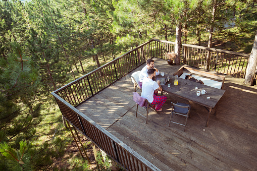 Two guys depicted from a high angle perspective, sitting on the balcony of a cottage in the woods. They're watching content on the laptop while still in their pajamas. Their dog is resting on the bench.