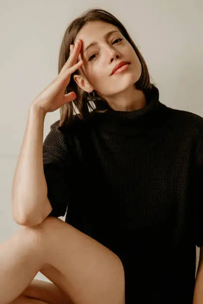 Portrait of a beautiful cozy woman in a black roll sweater sitting indoors. Being sensual and making simple poses. White background. Looking at the camera with one hand on her head.