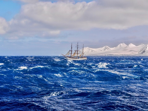 A traditional tall ship with reefed sails sailing through large waves with whitecaps, with mountainous land covered with snow and glaciers visible in the background.