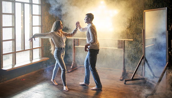 Two young people are practicing a Latin American dance. The man and the woman with long hair dressed in white shirts and blue jeans. The young woman is teaching the young man and showing him dance moves. Studio shooting in a dance classroom