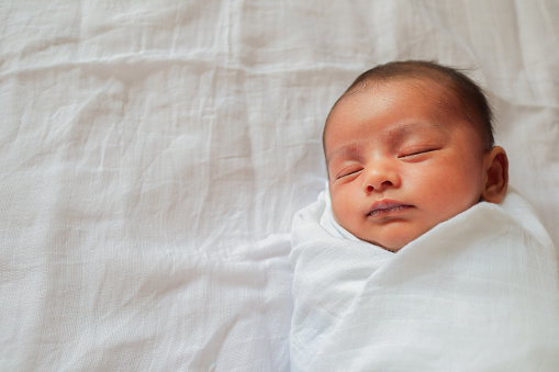 a one month old baby sleeping and swaddled in white cloth lying in white cloth background.
