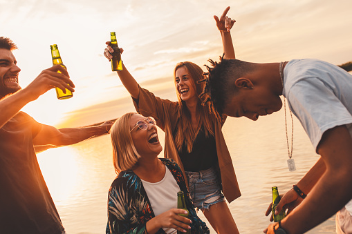 Group of young friends having fun drinking beer and dancing on pier by the lake at sunset.