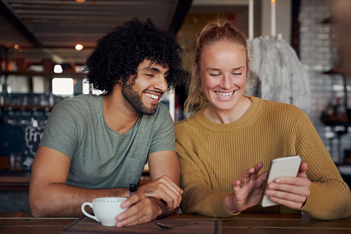 Cheerful young woman showing smartphone to boyfriend in modern cafe