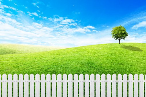 Long white wooden fence with Green grass meadow field and little hill with white clouds and blue sky in background.