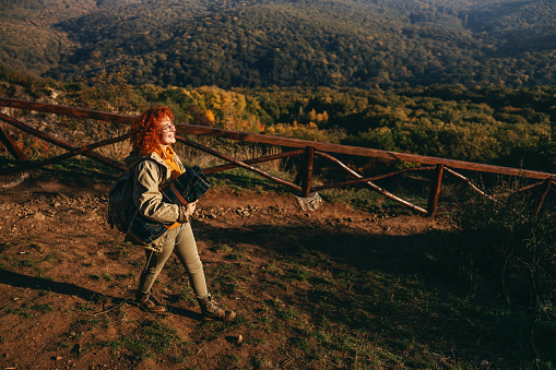 Full length of female ginger explorer having backpack on backs, holding blanket and walking in nature on a beautiful sunny autumn day.