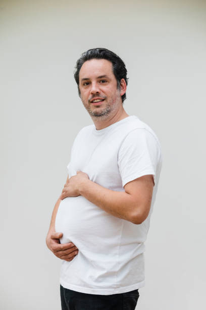 Look at my Belly Mature man smiling at the camera and sticking out his belly and touching his stomach pretending to be pregnant on a white background. pot belly stock pictures, royalty-free photos & images