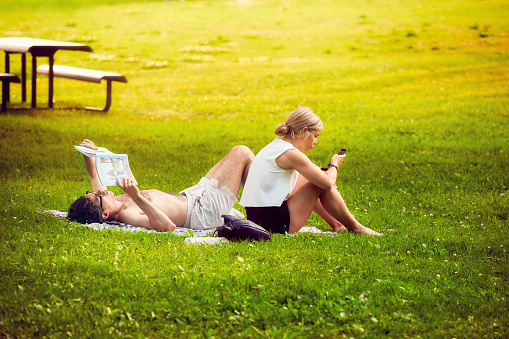 Montreal, Canada - June, 2018: Young Canadian couple sit at the green grasses of Mount Royal park in Montreal, Canada. Man reads a book and woman looks at the mobile phone