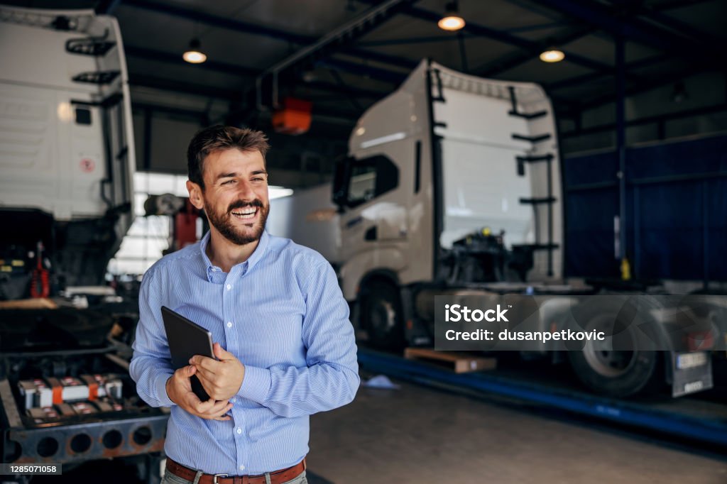 Smiling chief standing in auto park and holding tablet. In background are trucks. Firm for import and export. Freight Transportation Stock Photo